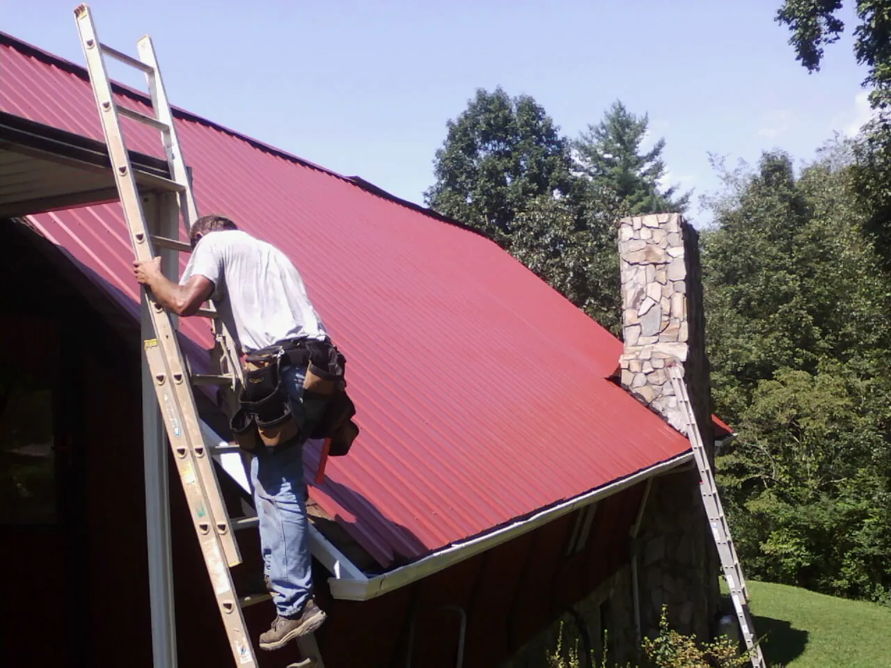Roofer on a ladder beside a red metal roof and stone chimney — Soffit & Fascia Repair by Roofing Brothers in Kirkland